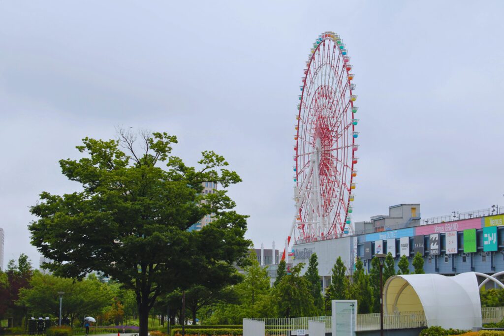 A tall red and white Ferris wheel beside a modern shopping complex on a cloudy day.