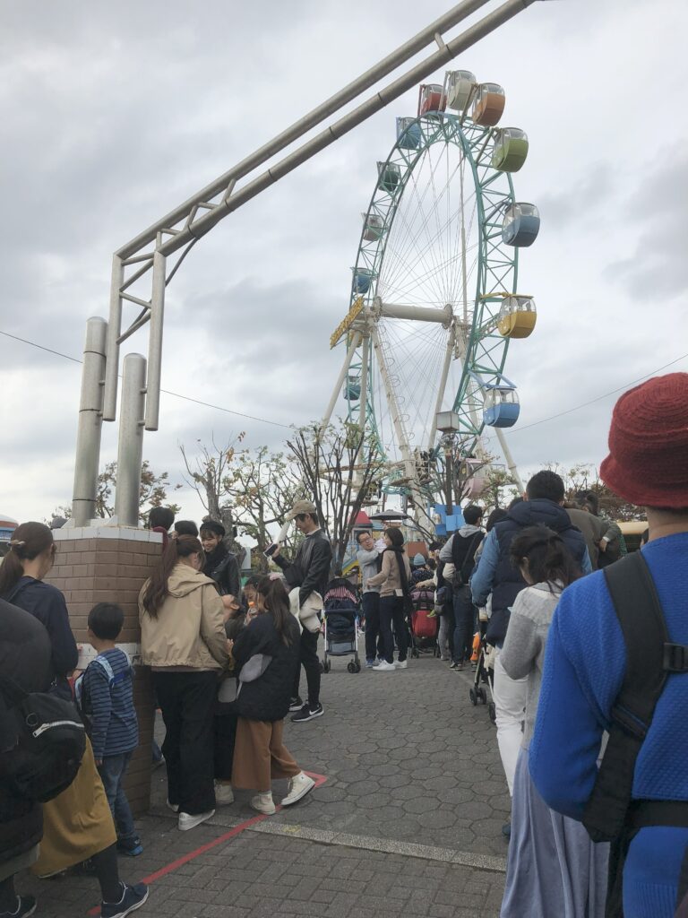 People lining up outside a classic amusement park entrance with visible trees and tram lines nearby.