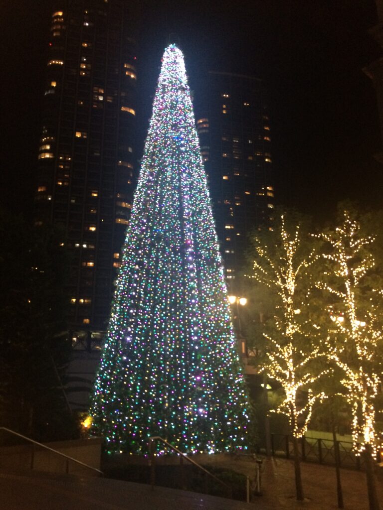 A towering Christmas tree covered in colorful lights, surrounded by city skyscrapers and lit trees.