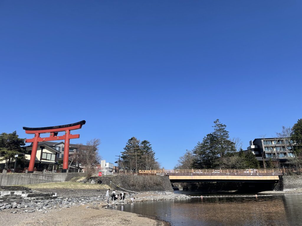 View of a red torii gate and a small pedestrian bridge by the riverbank in Nikko with a blue sky overhead.