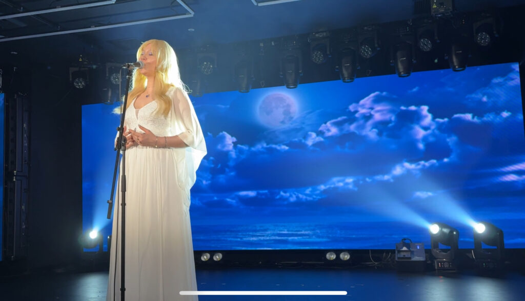Woman in a white gown singing onstage with a moonlit background