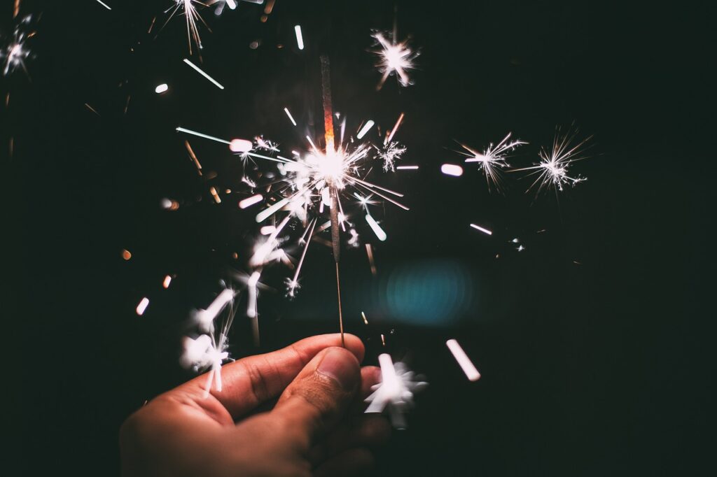 A hand holding a lit sparkler at night with bright sparks flying in the dark.