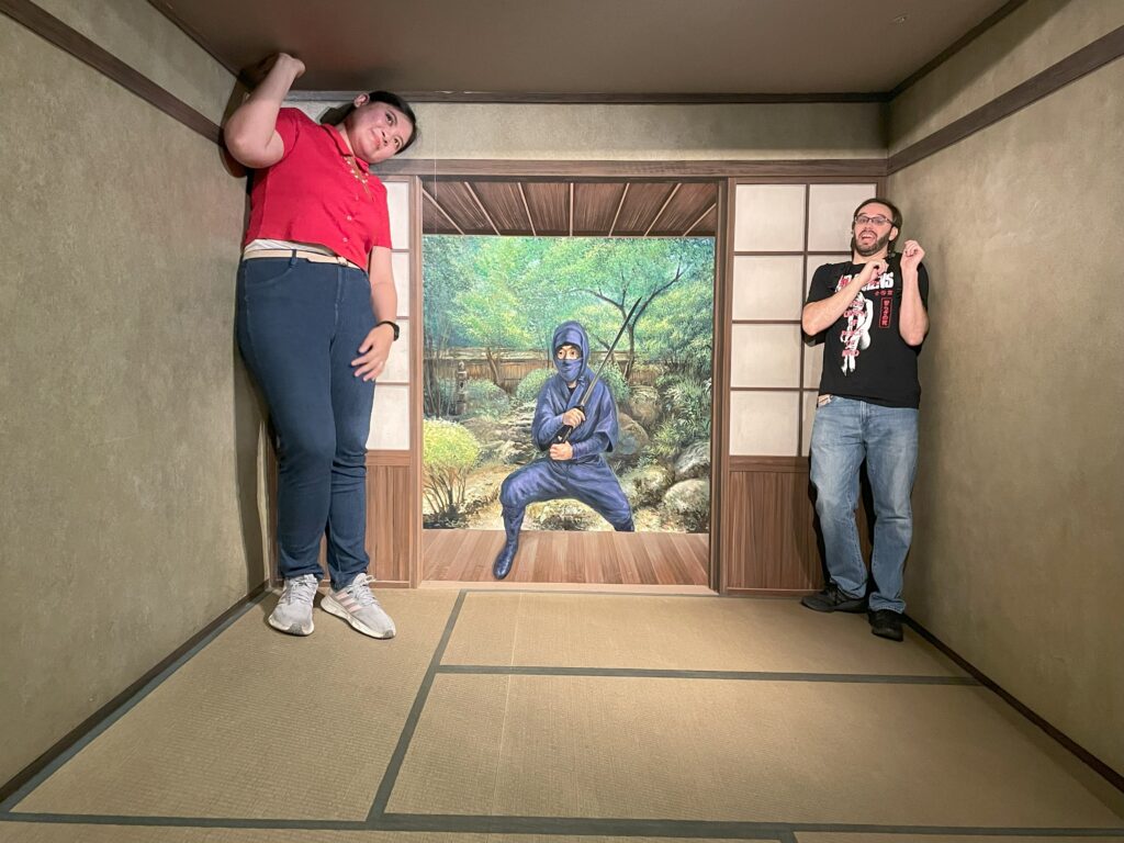 A woman appears to be a giant leaning on the ceiling while a man looks tiny in a traditional Japanese room illusion.