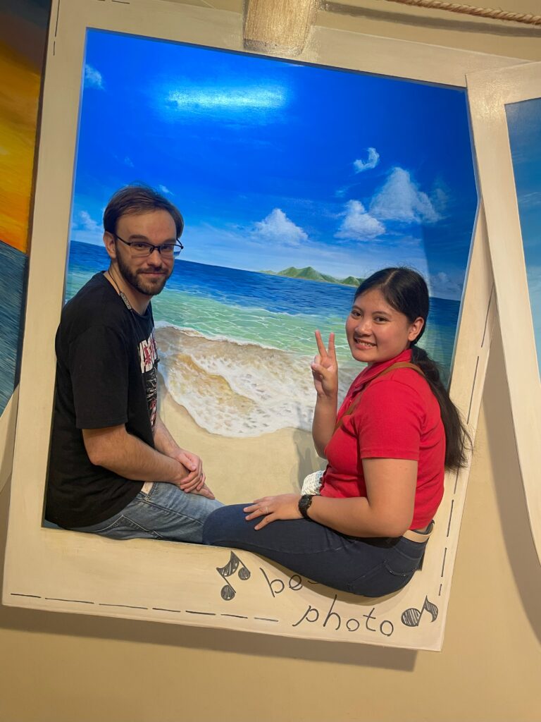 Two people sitting inside a beach-themed illusion frame at Tokyo Trick Museum.