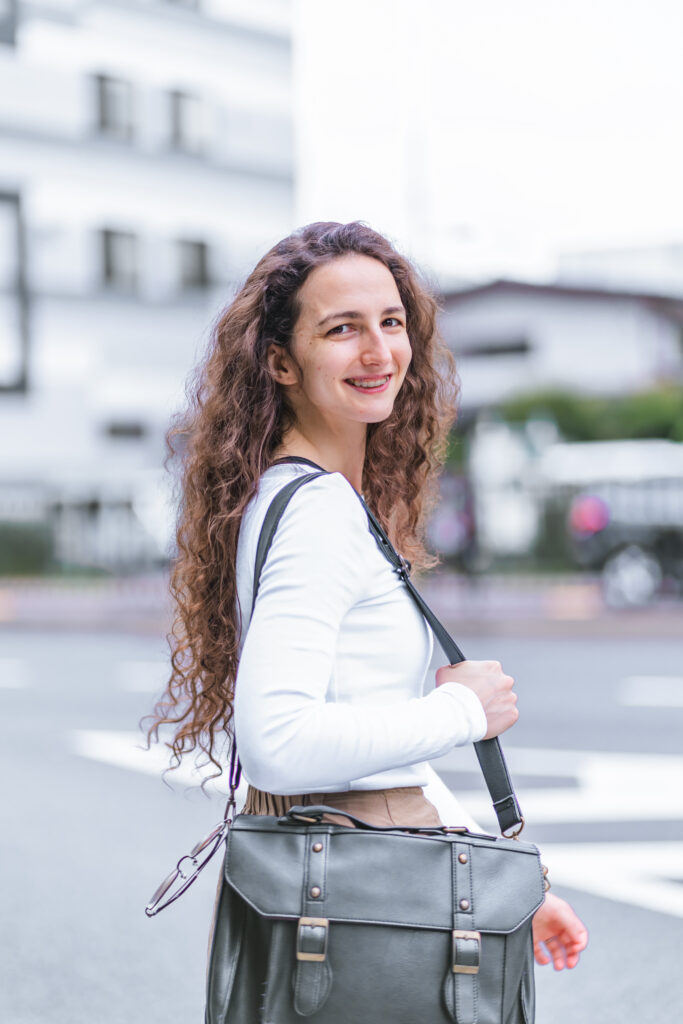 Mar Exposito in a white long-sleeve top, turning around and smiling while holding a black bag, walking in Tokyo.