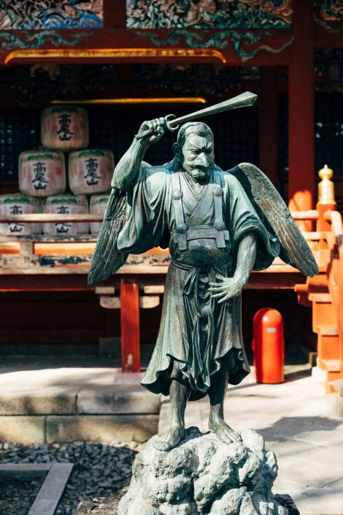 A bronze statue of a tengu with wings and a raised sword, standing guard at a shrine.