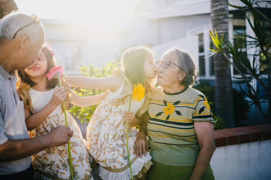 Granddaughter kisses grandmother on the cheek while holding a flower, surrounded by family in warm sunlight.