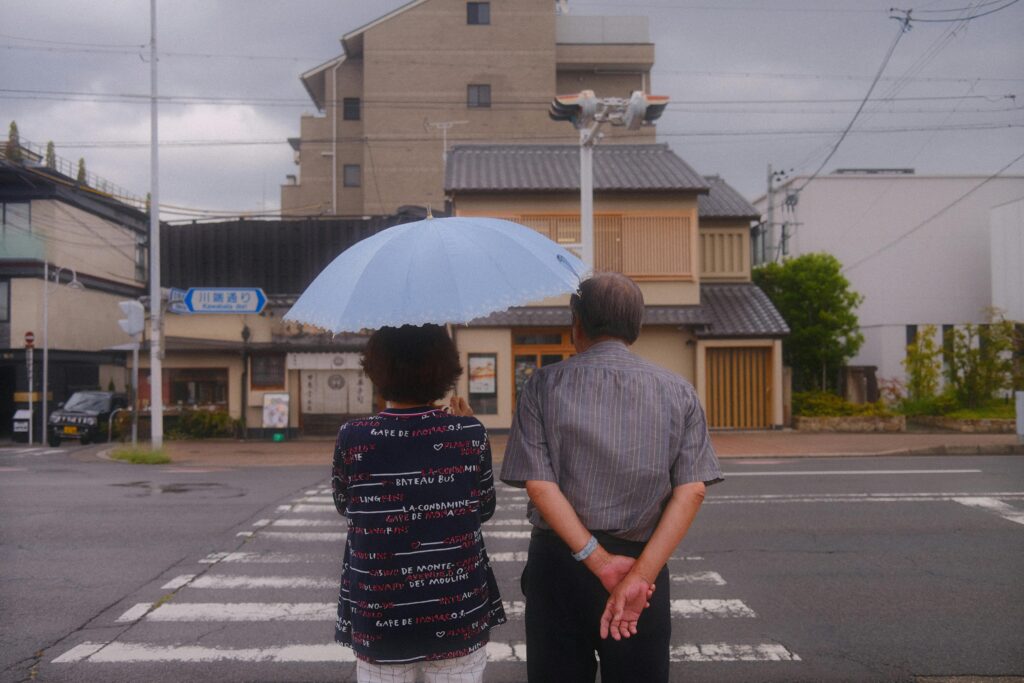 Elderly couple standing under a blue umbrella on a crosswalk in a quiet Japanese neighborhood.
