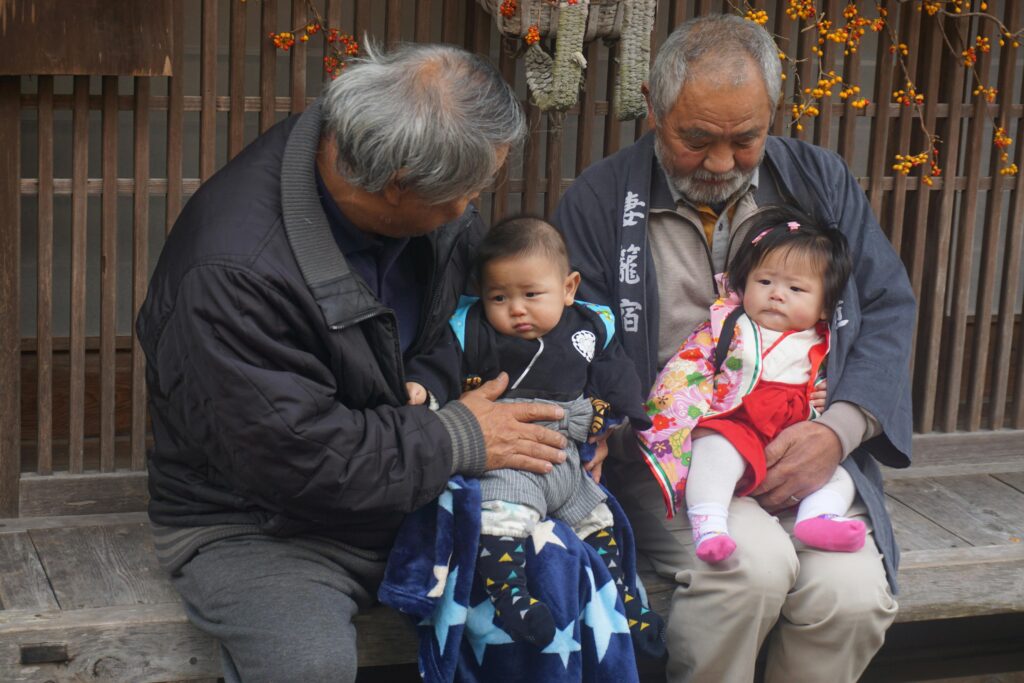 Two elderly men holding babies dressed in traditional clothing while sitting on a wooden bench.