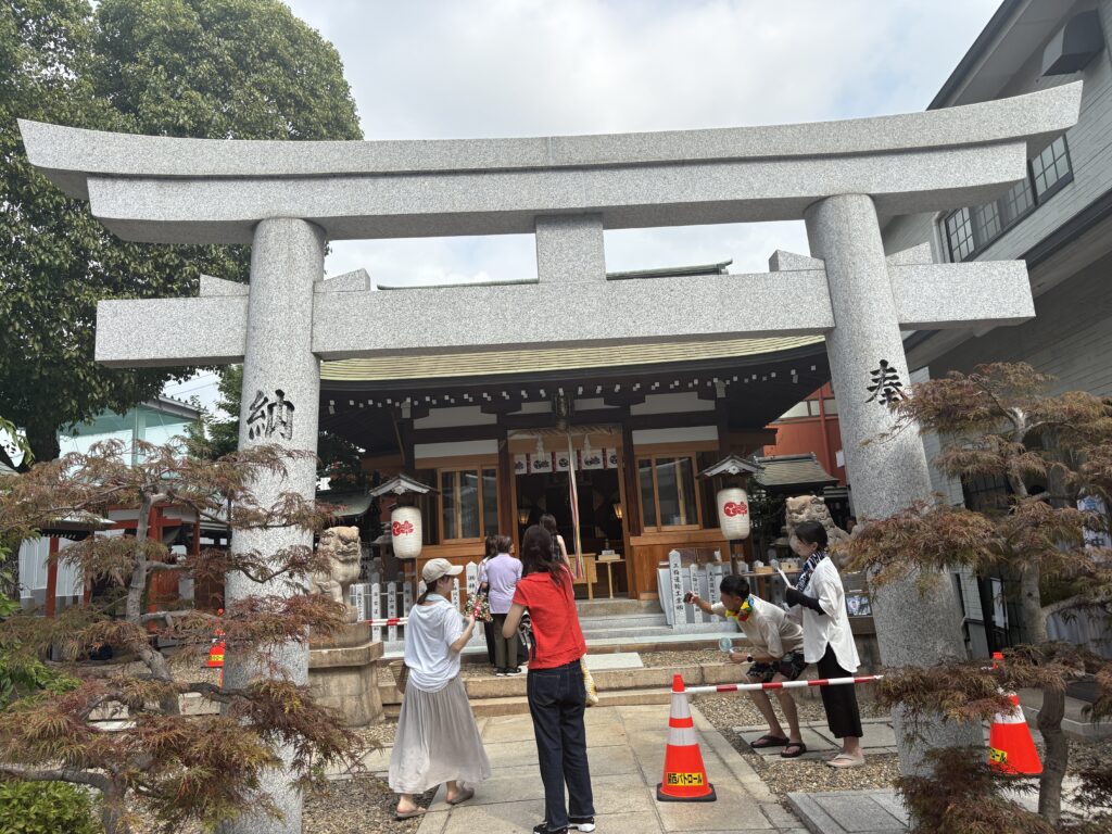 A hand holding a red embroidered omamori charm in front of the heart-shaped tree at Wakinohama Shrine.
