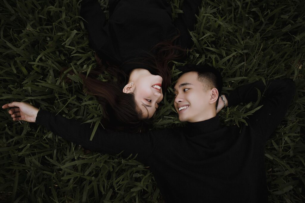 A couple lying on the grass facing each other and smiling while wearing matching black sweaters.
