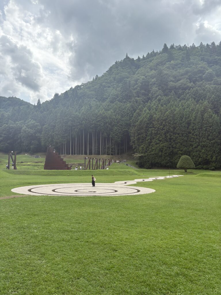 Wide view of Morou Art Forest with a circular stone pattern and sculptures against a forested mountain backdrop.