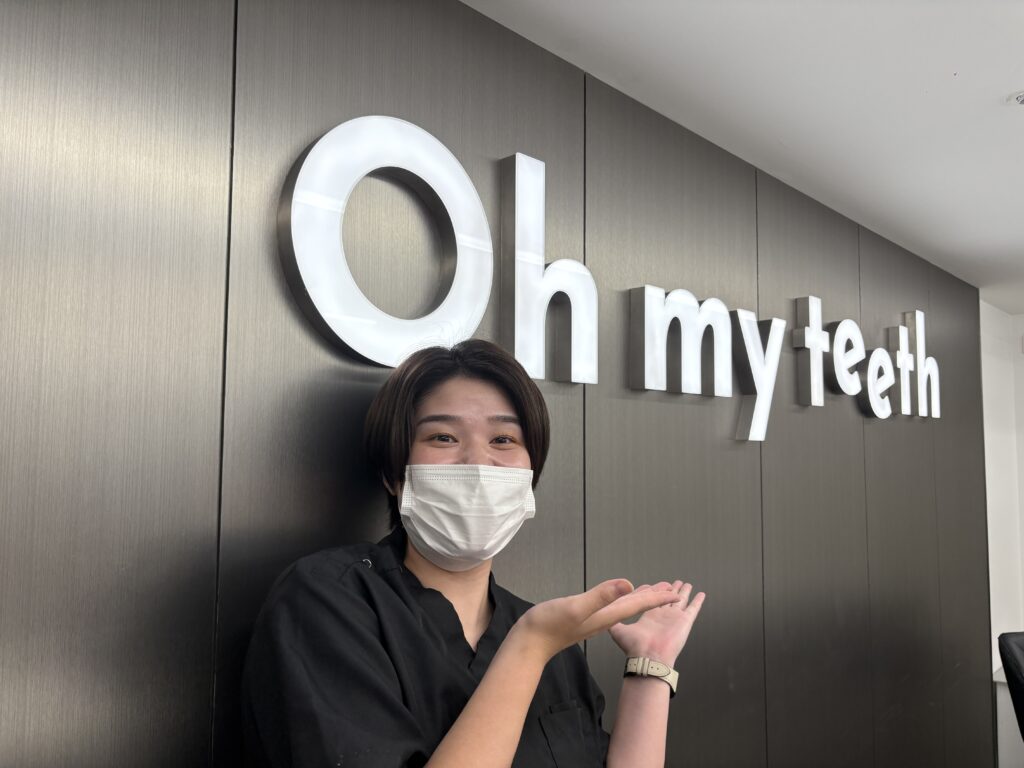 Female dental staff member wearing a face mask and black uniform standing in front of the illuminated “Oh my teeth” clinic sign, smiling and gesturing with open hands.