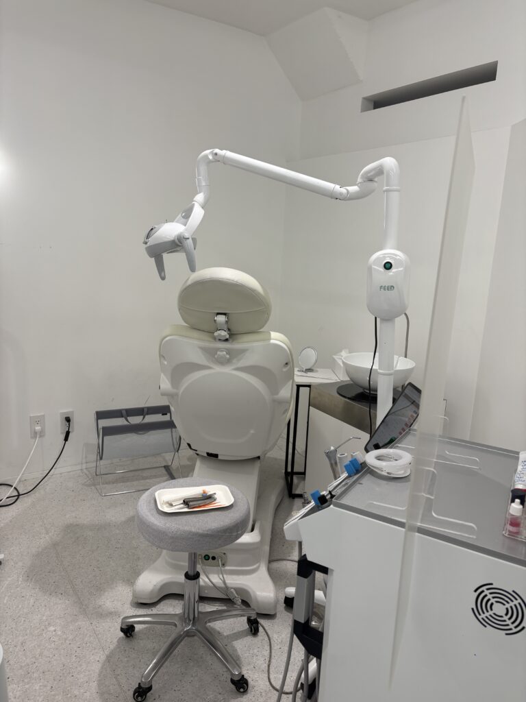 Minimalist dental treatment room featuring a reclining white dental chair, stool, instruments on a tray, and clinical equipment under bright lighting.