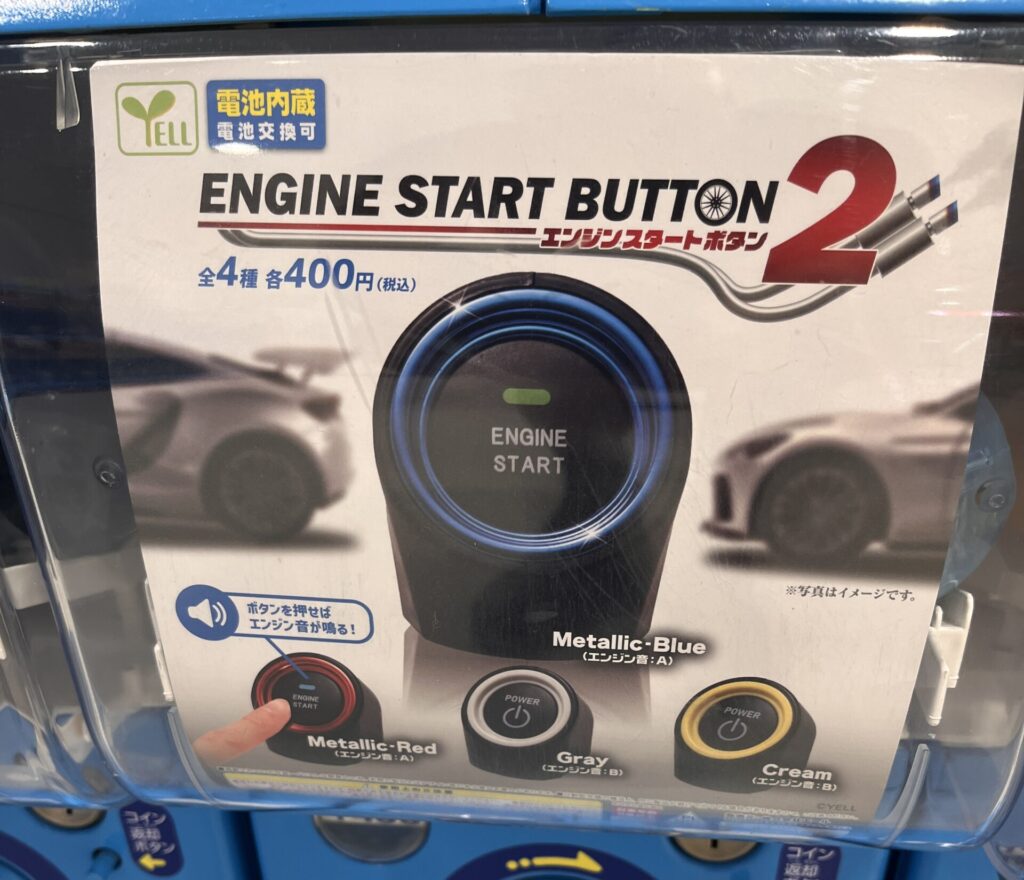 Close-up of a gachapon machine featuring an “Engine Start Button” toy in four colors — metallic blue, red, gray, and cream — each costing 400 yen.