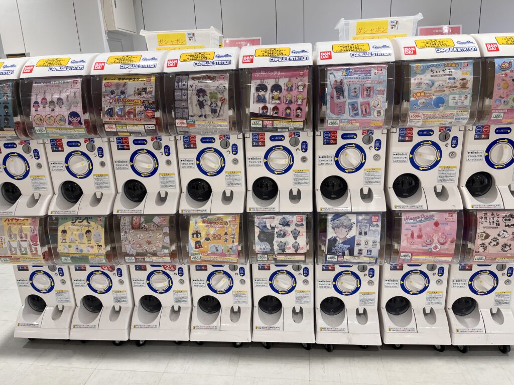 A full row of gachapon machines in a shopping center, each offering different capsule toys such as anime characters, mascots, and accessories.