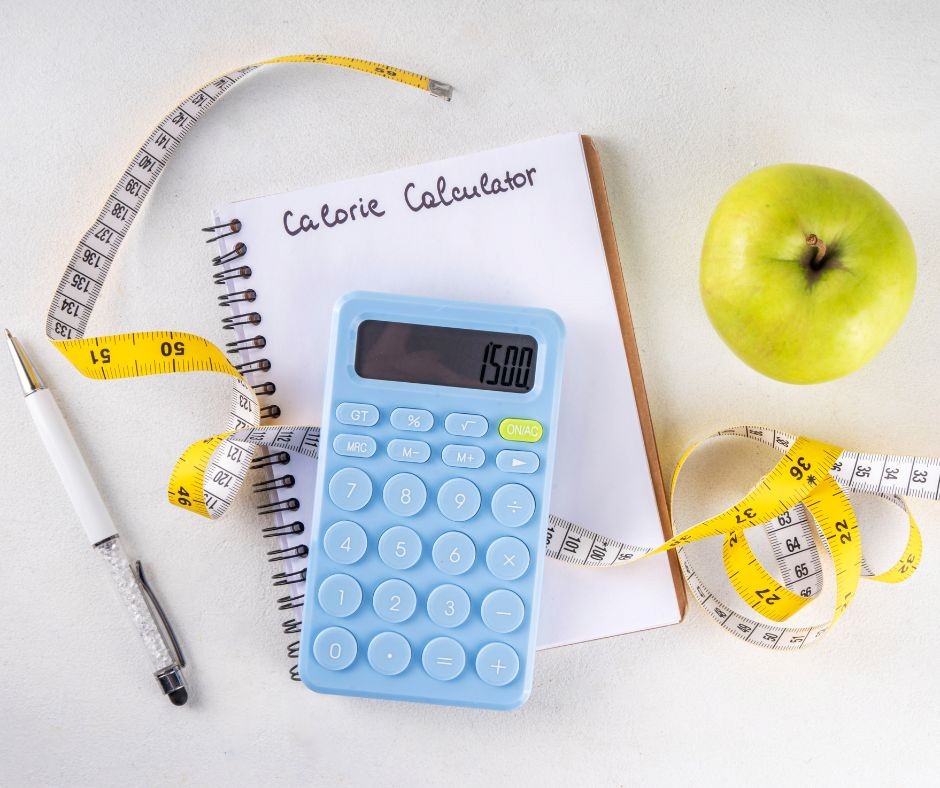 Calorie calculator setup with a notebook, pen, tape measure, green apple, and blue calculator on a white table.