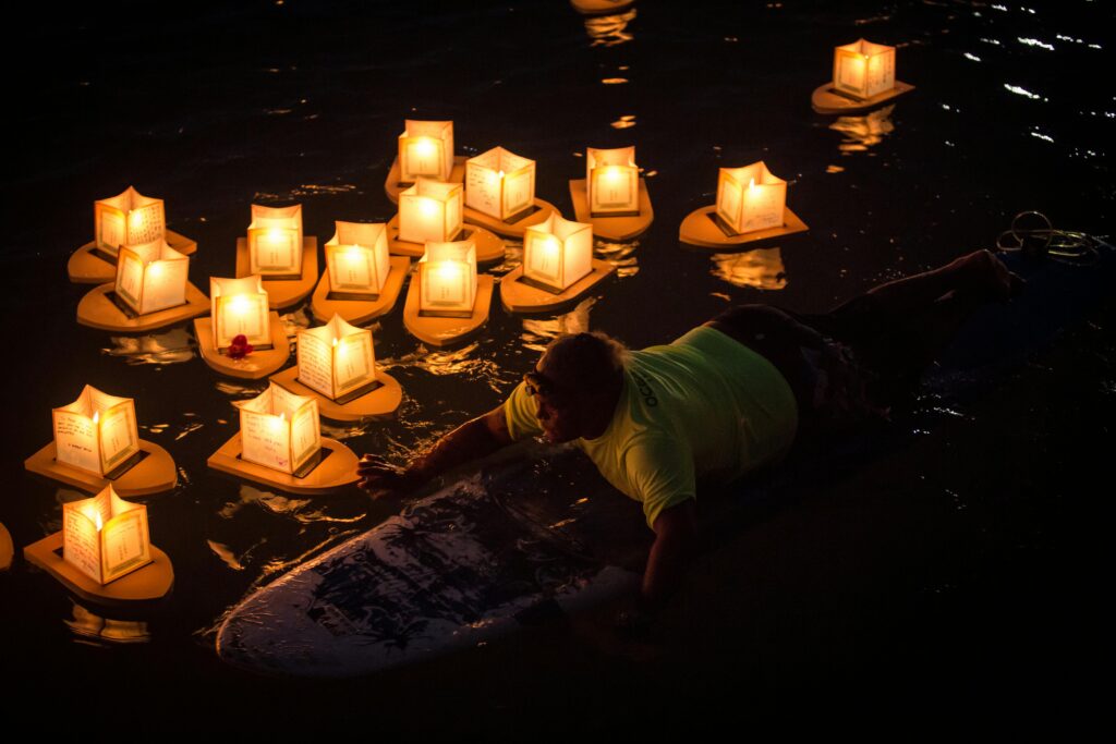  A man on a surfboard in the water surrounded by glowing paper lanterns during a nighttime lantern floating ceremony.