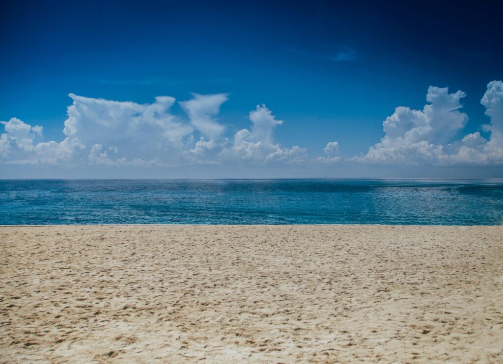 A clear sunny day at the beach with golden sand in the foreground and a calm blue ocean stretching to the horizon under fluffy white clouds.