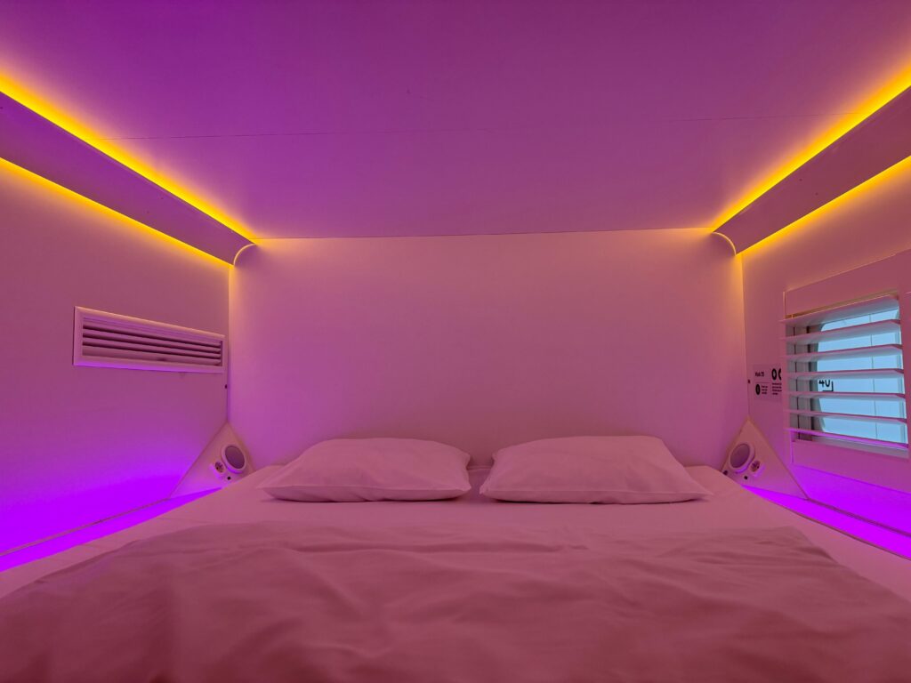 Interior of a modern capsule hotel pod with white bedding and ambient purple and yellow LED lighting along the walls and ceiling.