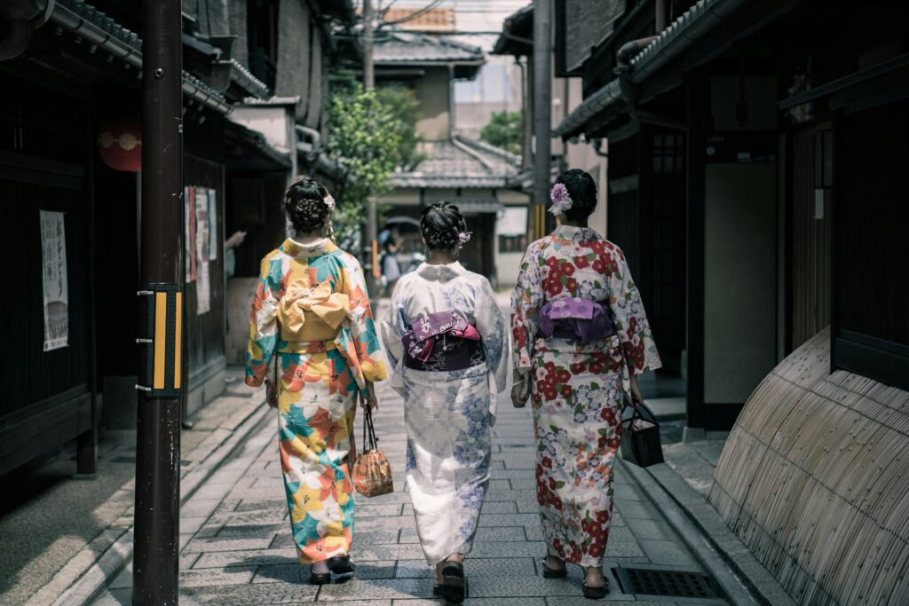 Three women dressed in colorful yukata walk down a narrow traditional Japanese street, each with a decorative obi tied at the back.