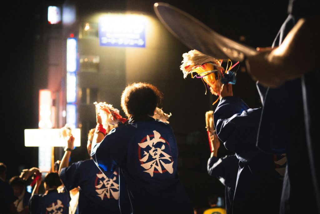 A group of dancers wearing traditional blue happi coats perform at night during a Japanese festival, holding colorful