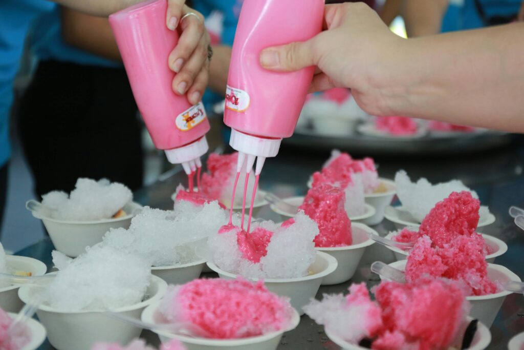 Close-up of hands pouring bright pink syrup over cups of shaved ice at a festival or summer event.