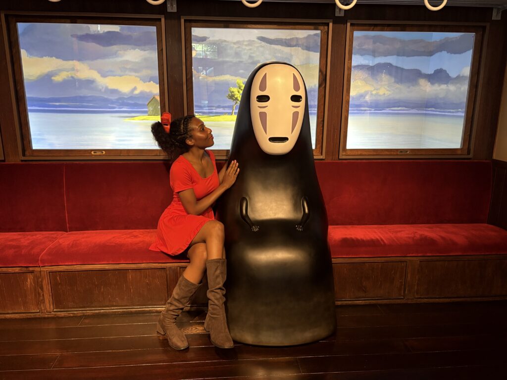 Visitor in a red dress posing beside No-Face on the iconic red train seat from Spirited Away.