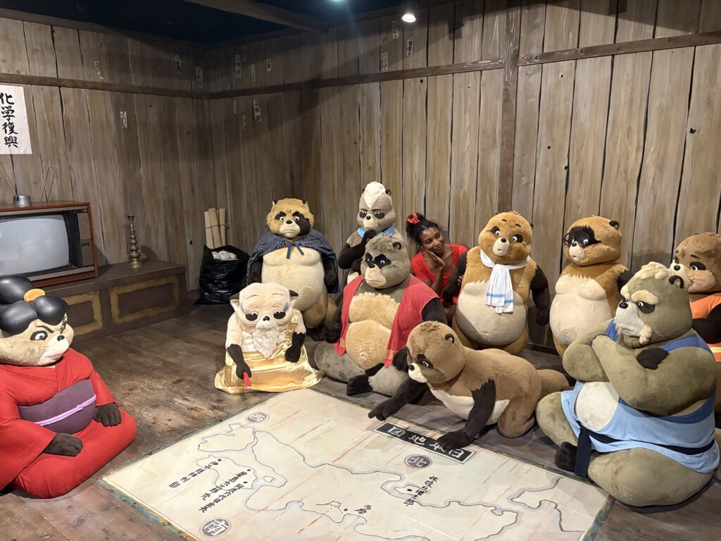 Life-sized raccoon dog figures from Pom Poko gathered around a map inside Mononoke Village, with a visitor in a red dress posing among them.
