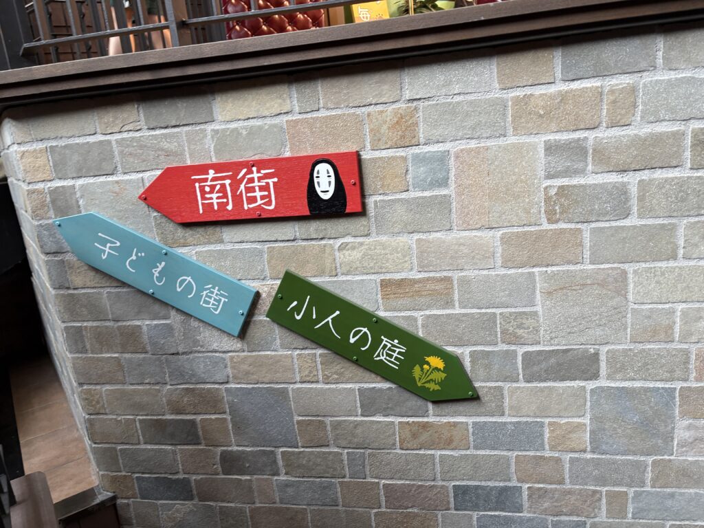 Wooden direction signs on a stone wall inside Studio Ghibli Park, including an arrow with No-Face from Spirited Away.