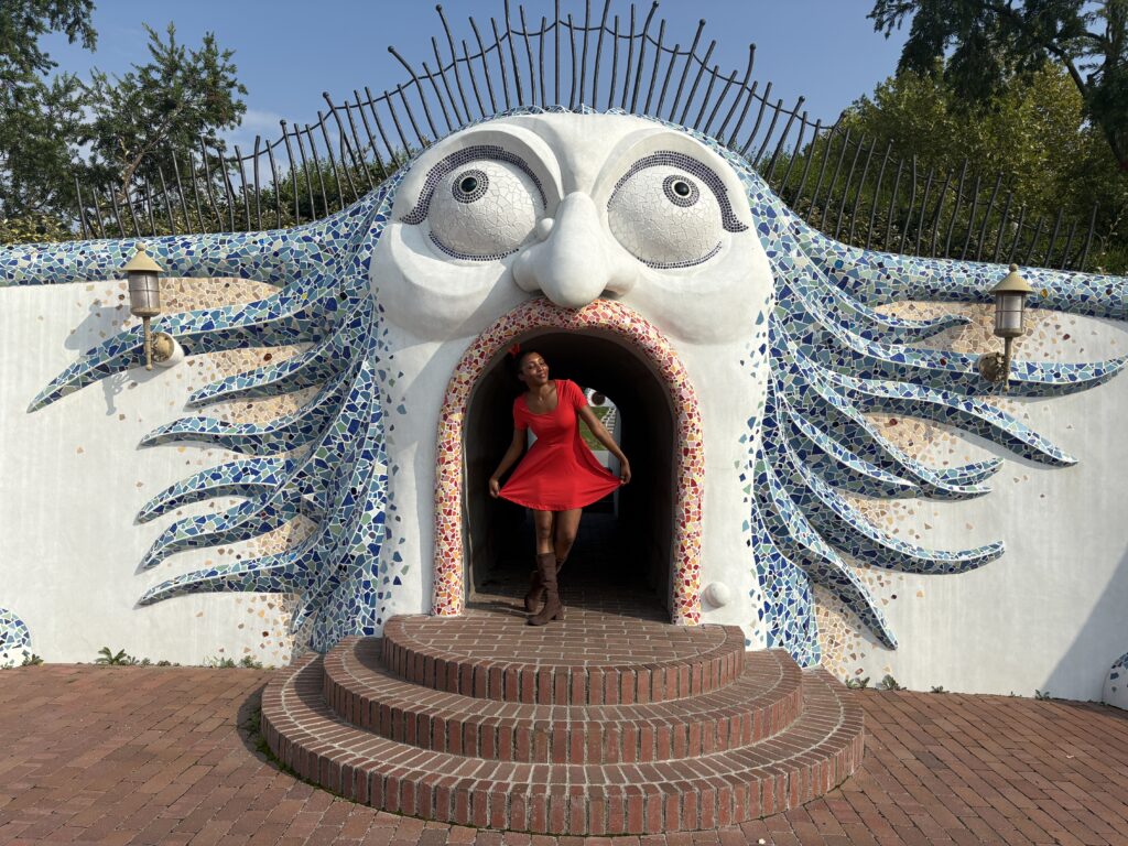 Visitor in a red dress stepping out of a whimsical tiled face-shaped entrance with mosaic blue hair at Ghibli Park.