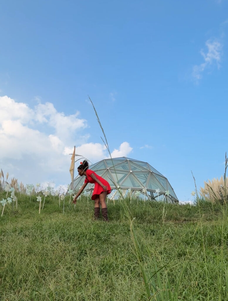 Visitor in a red dress standing on a grassy hill near a dome-shaped glass structure, pointing toward the ground.