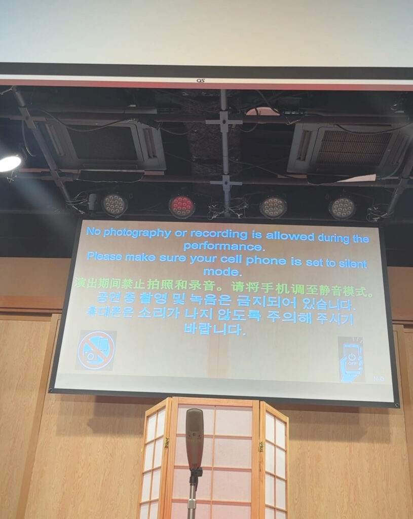 Screen inside the theatre showing “No photography or recording is allowed” in multiple languages with a microphone in front.