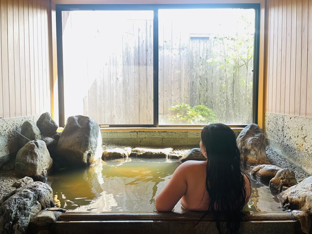 A woman with long black hair relaxing in a private rock bath onsen in Chiba, with sunlight streaming through a large window overlooking a small garden.