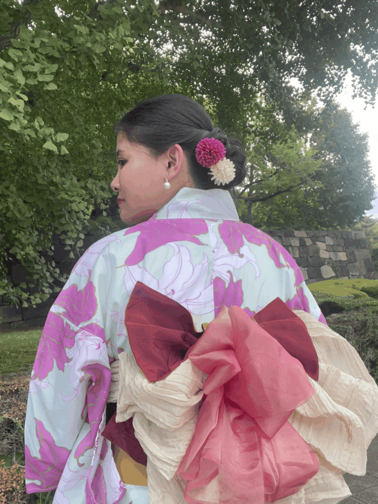 Back view of a woman in a pink floral yukata with a decorative layered obi, standing outdoors with stone walls and greenery behind her.