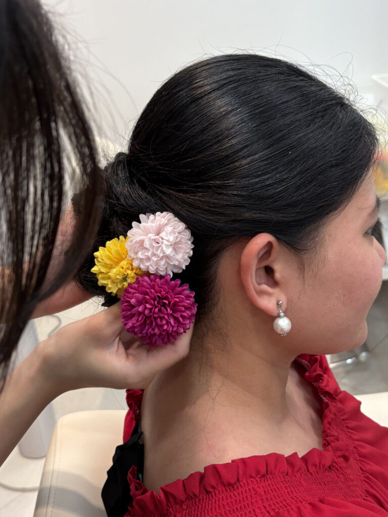 Hairstylist attaching colorful floral hair accessories (pink, yellow, white) to a neatly styled bun.