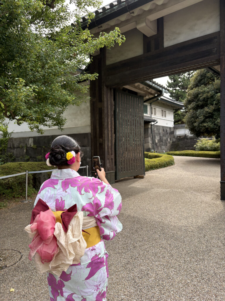 Woman in a floral yukata taking a photo with her phone near the entrance gate of the Imperial Palace, her elaborate obi visible from behind.