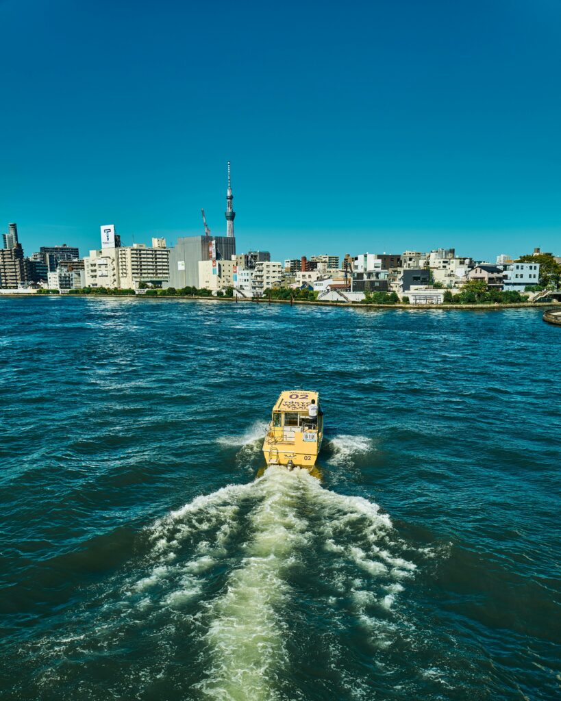 A bright yellow taxi boat sailing across Tokyo Bay, leaving a white wake behind, with the city skyline ahead.
