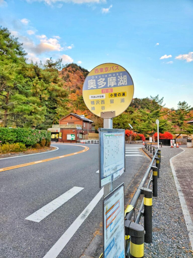 A road-side bus stop sign reading “Okutamako” with autumn trees and mountains in the distance.