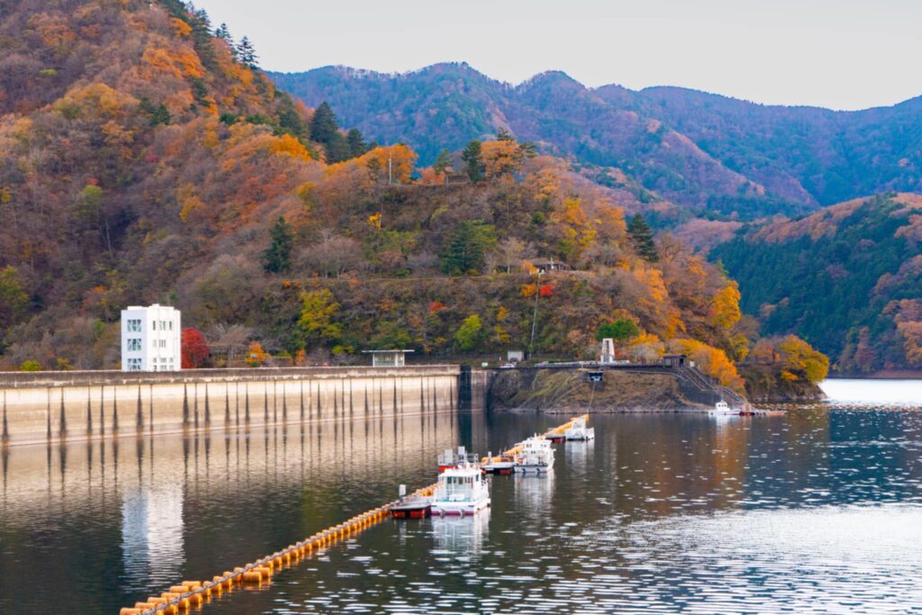 A scenic view of Ogouchi Dam with boats floating on the calm lake, backed by mountains covered in fall colors.