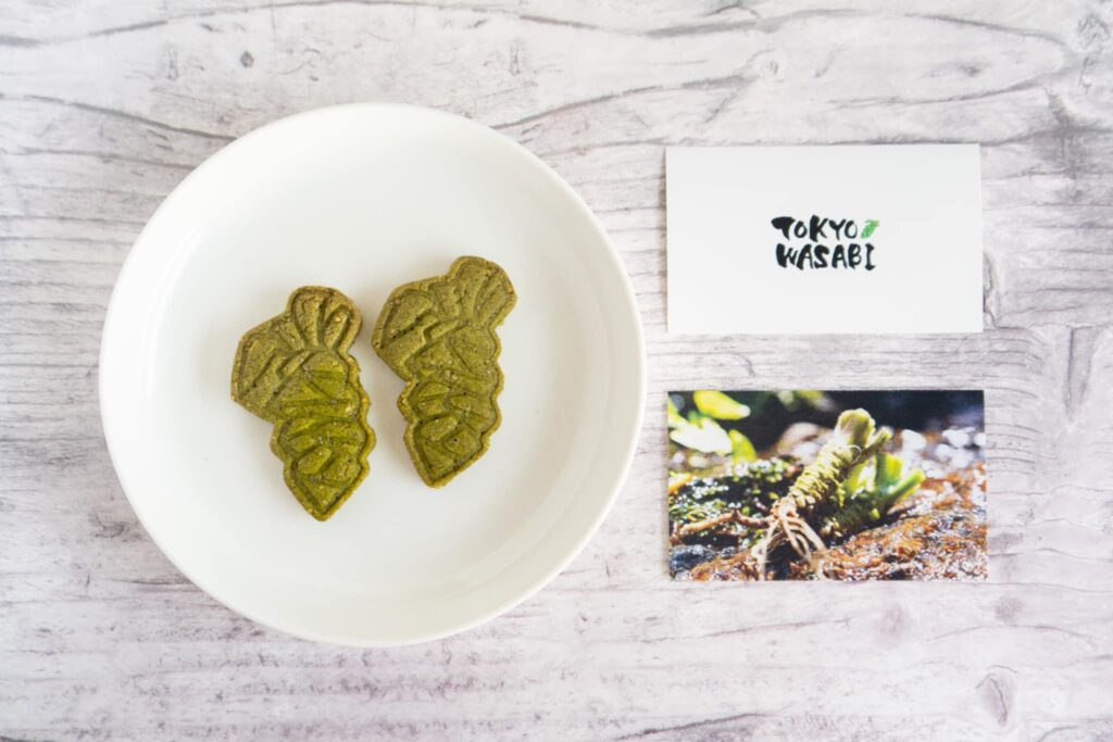 Two green wasabi-shaped cookies on a white plate with promotional cards in the background.