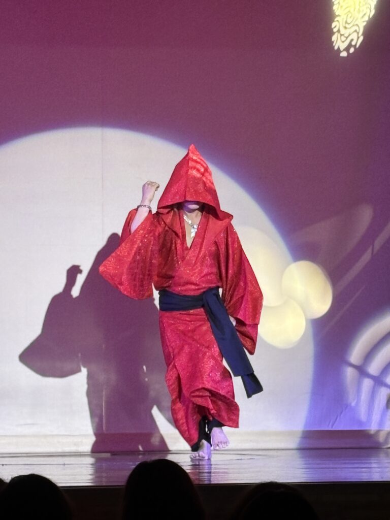 A performer dressed in a bright red kimono with a hood on stage, striking a dramatic pose under colorful lighting.