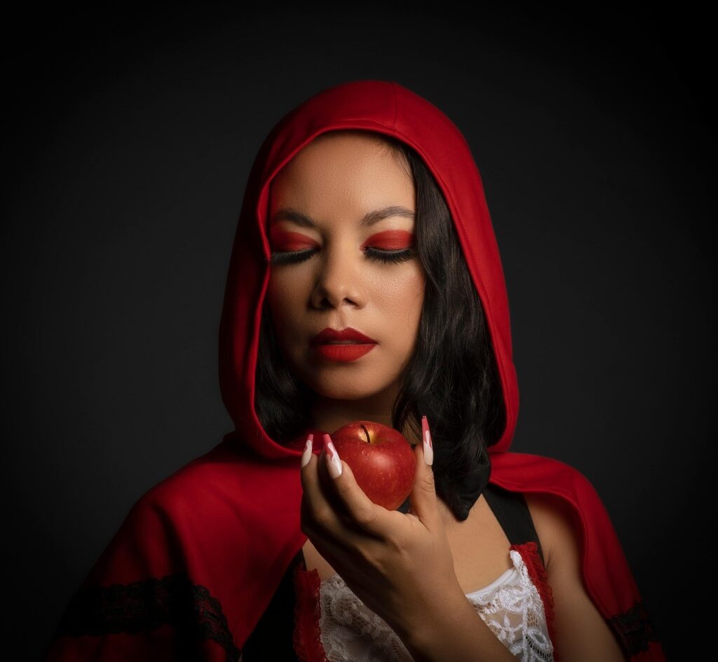 Woman dressed as Little Red Riding Hood holding a red apple with eyes closed, wearing bold red makeup and cape against a dark background.