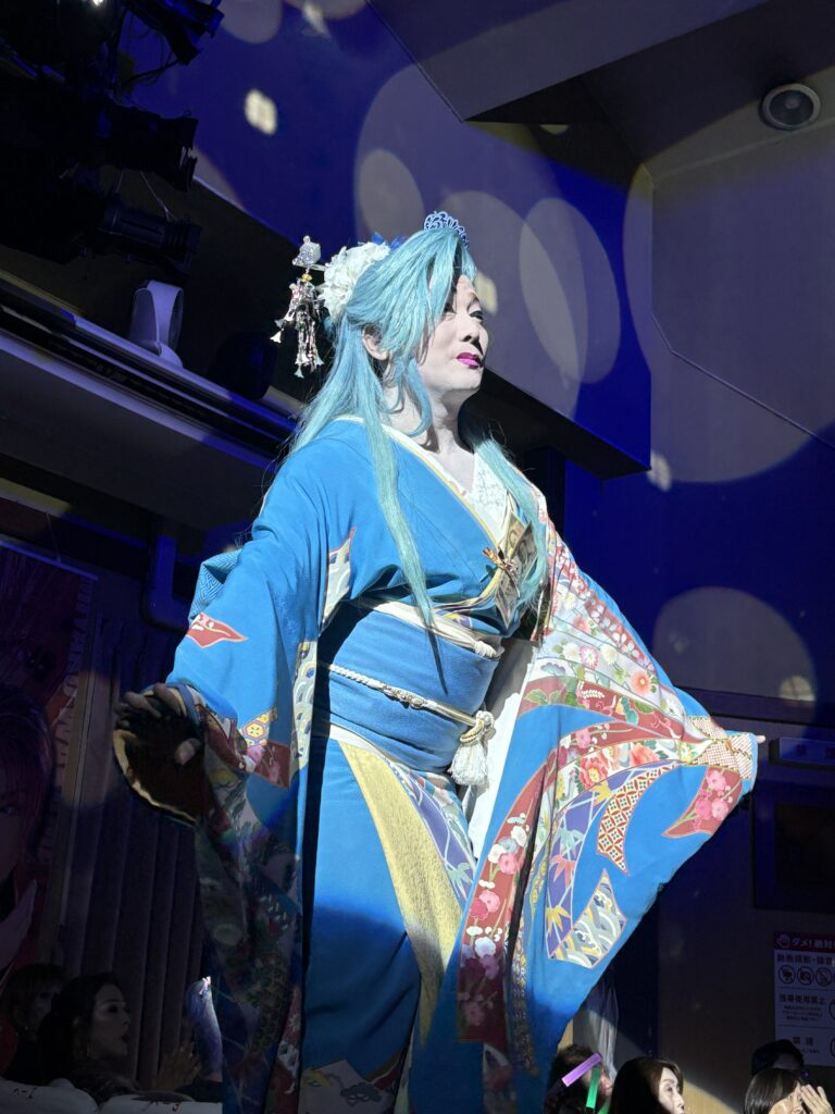A performer in a blue kimono and matching wig with elaborate hair accessories performing dramatically under stage lighting.