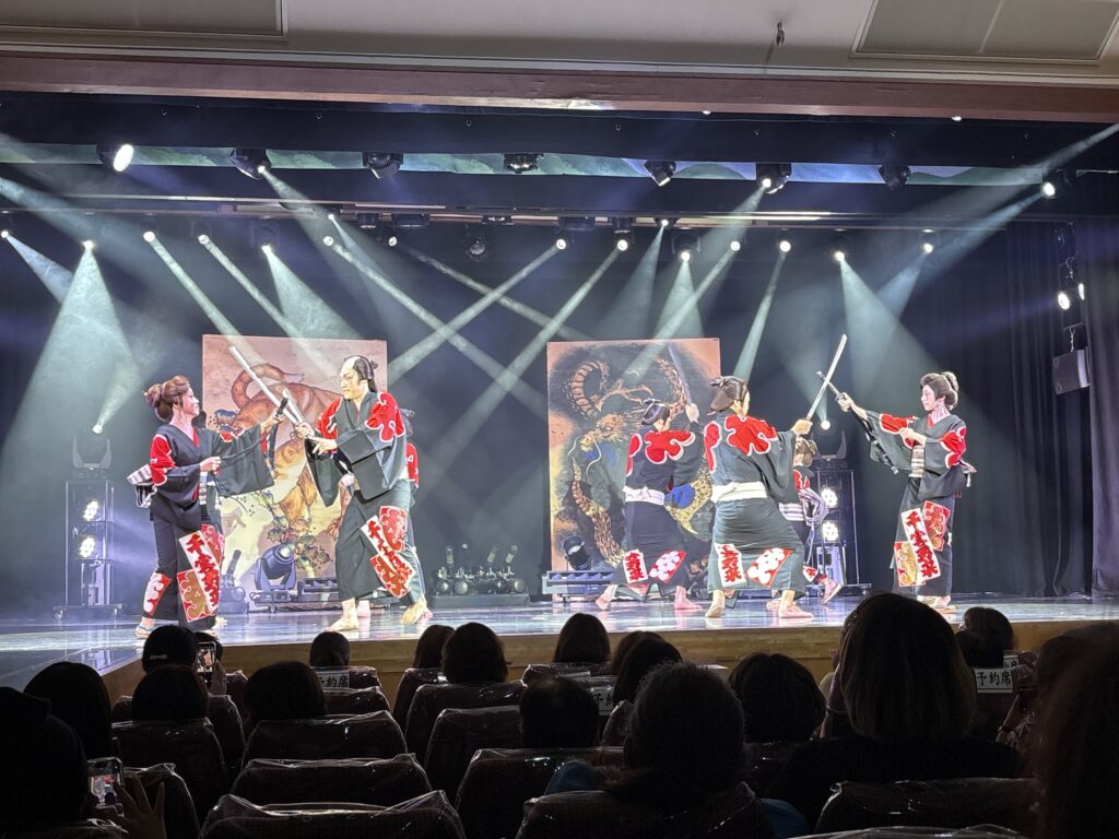 Multiple Taishuu Theatre performers dressed in matching black and red costumes perform a choreographed sword-fighting scene on stage, illuminated by dramatic lighting.
