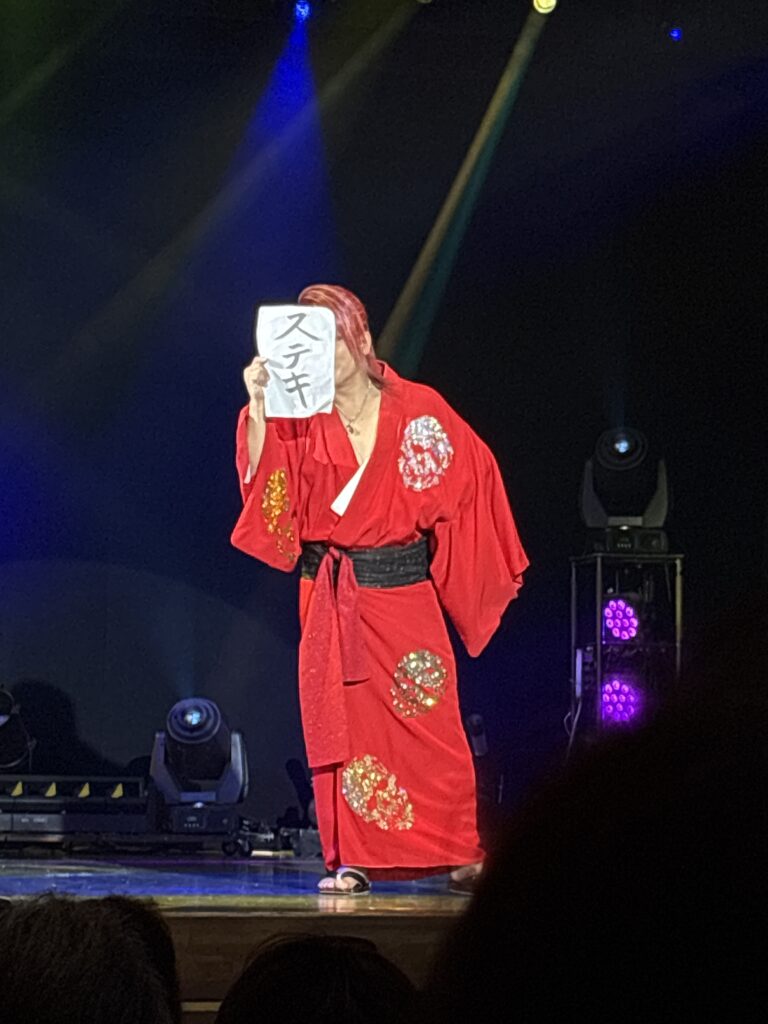 A Taishuu Theatre actor in a red kimono holds up a paper sign with Japanese writing (“ステキ,” meaning “wonderful”) during a comedic stage performance.