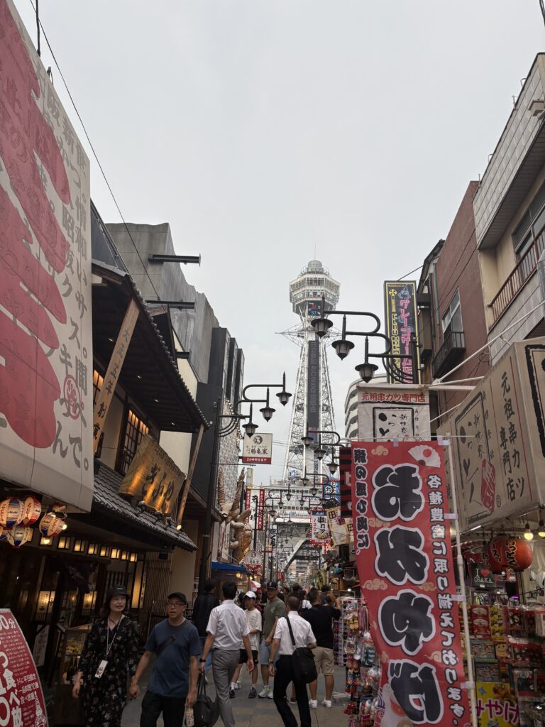 A busy street in Osaka’s Shinsekai district with Tsutenkaku Tower in the background, colorful shop signs, and people walking under lanterns and banners.