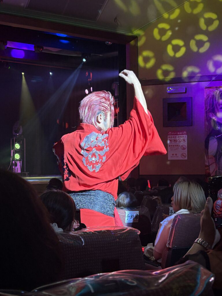 A performer with pink hair wearing a red kimono dances in front of an audience under spotlights inside the theater.