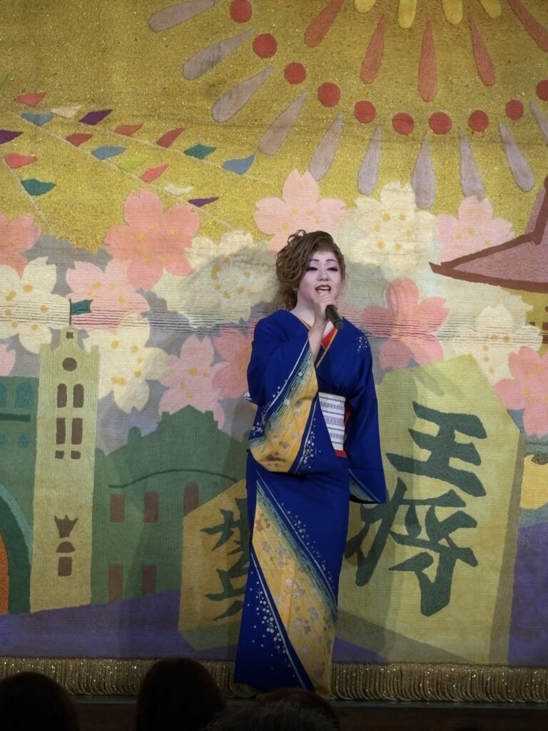 A female performer in a blue kimono sings on stage with a colorful cherry blossom and sun-themed backdrop.