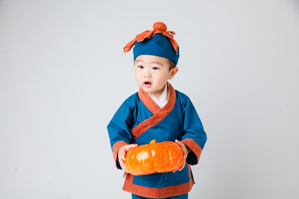 Young child in blue and orange traditional outfit holding a small pumpkin against a white background.